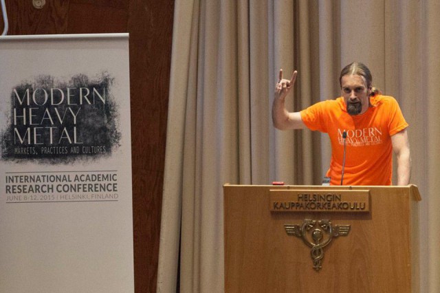 Toni-Matti Karjalainen avasi konferessin. Totta helvetissä puhujakopissa nostetaan pikkurilli ja etusormi ilmaan lajiuskollisuuden merkiksi.